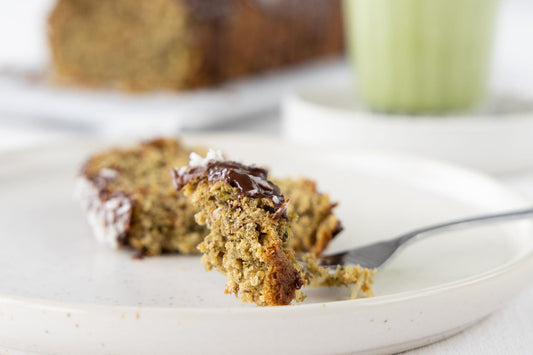 Ein Stückchen Matcha-Kiwikuchen mit Schokoguss auf einer Gabel. Das angeschnittene Stück des Kastenkuchens liegt dahinter. Nach weiter im HIntergrund steht ein Matcha-Latte in einem Glas und der Rest des Kuchens.