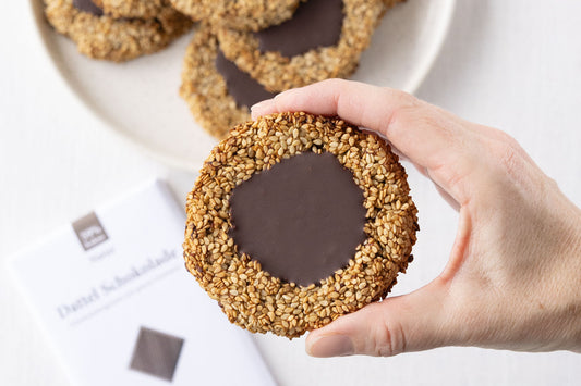 Sesam-Cookie mit Schokoladenfüllung zwischen zwei Fingern gehalten. Im Hintergrund sind weitere Cookies und eine Tafel DattelScokolade zu sehen